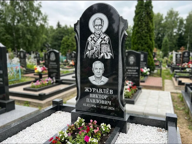 Памятник с гравировкой иконы: стела с образом в полный рост на обороте — гравировка образа Матроны Московской, низкие кусты спиреи в цвету, работа «Лебедева» в Иркутске
